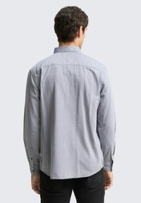 Chemise à carreaux grise avec manches longues, ourlet arrondi et poignets boutonnés. Le tissu présente un motif texturé et léger.