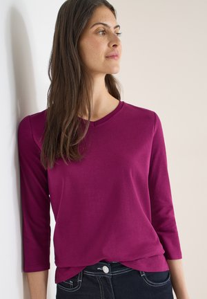 Fuchsia langærmet top med rund halsudskæring og blød tekstur, kombineret med mørke jeans, lidt stoppet ind ved taljen. Enkelt design.