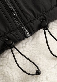 Black quilted jacket featuring a central zipper and adjustable drawstrings. The interior has a fluffy white texture, enhancing warmth.