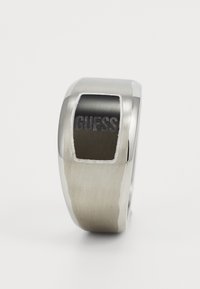 Bague en acier inoxydable avec une finition argentée brossée et un accent noir poli. Gravé "GUESS" à l'avant dans une couleur contrastante.