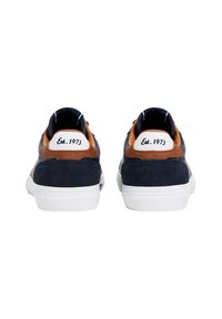 Tengerészkék sportcipők barna szarvasbőr részletekkel, fehér gumitalppal és egy sarokfüllel, amely fekete felirattal "Est. 1973" szerepel.