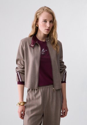 Mujer que lleva una chaqueta marrón con rayas finas y cremallera, pantalones a juego y una camisa granate debajo, con pulseras doradas en la muñeca.