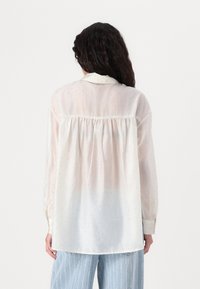 Gjennomsiktig hvit bluse med en svak gradient, prydet med små glitrende prikker, med gathered bakstykket og lange ermer med knapper ved håndleddene.
