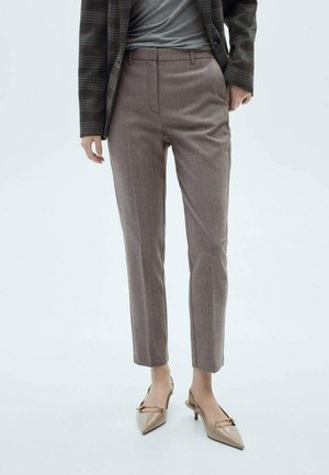 Persoon die op maat gemaakte bruine cropped broeken, een geruite blazer, een grijze top en beige puntige lage hakken draagt, staand op een lichte achtergrond.