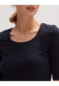 Femme à la peau claire et aux lèvres roses portant un t-shirt noir à col rond et manches courtes sur un fond uni.