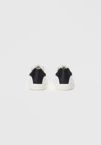 White sneakers featuring a black textured back panel, smooth white sides, and a flat rubber sole. Minimalist design with subtle branding.