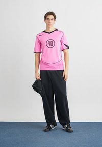 Κοντομάνικη φανέλα Nike σε έντονο ροζ με μαύρες λεπτομέρειες, με κυκλικό γραφικό '90', συνδυασμένη με χαλαρό μαύρο παντελόνι και μαύρο καπέλο.