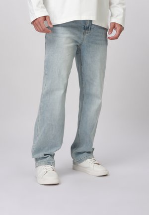 Mees, kes kannab helehalli sirgelõikelisi teksaseid, valgeid tossusid ja valget pika varrukaga särki, seisab helehalli taustal.