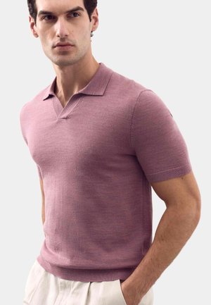 Uomo che indossa una polo rosa polvere a maniche corte con un colletto sottile, abbinata a pantaloni beige chiaro, in piedi con le mani in tasca.