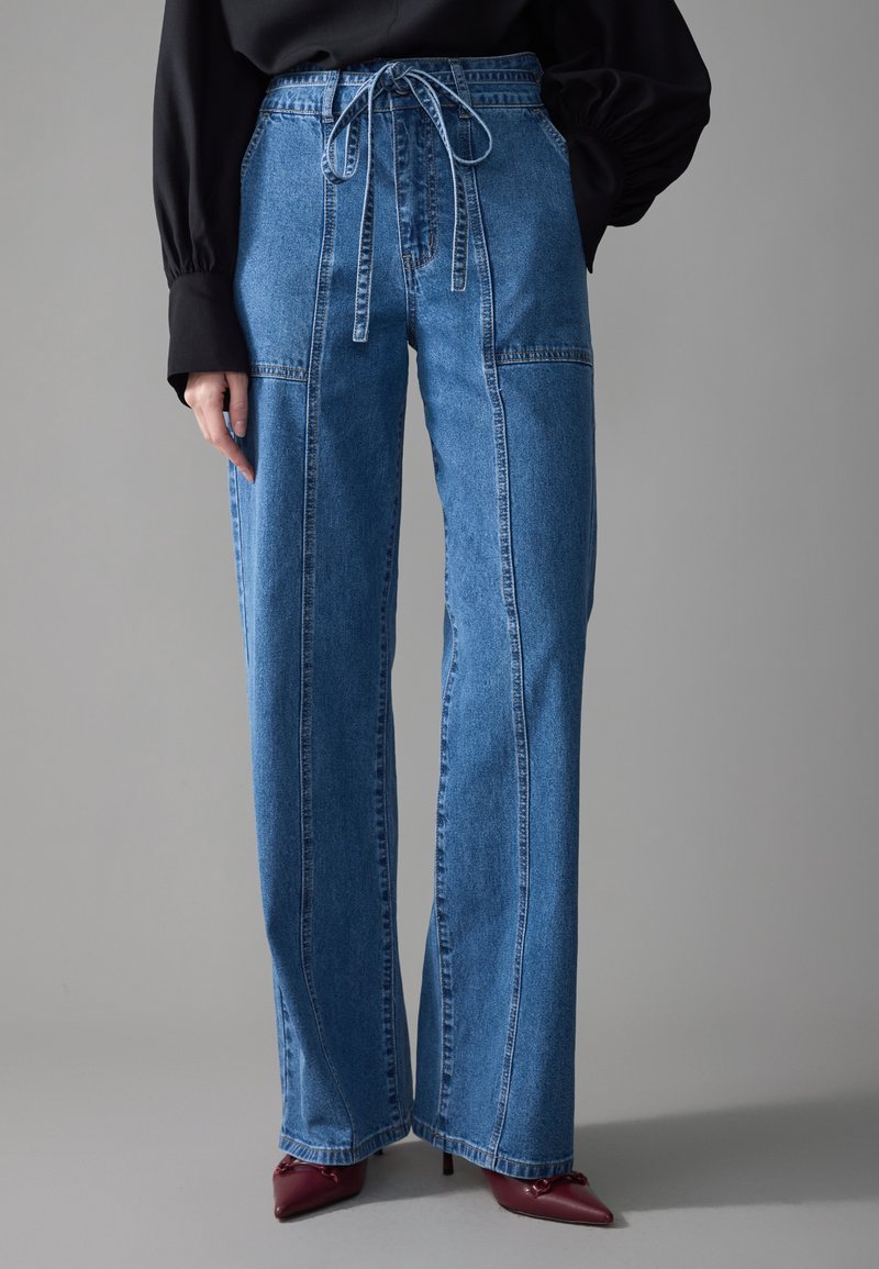 Bredde ben blå denim-jeans med høy midje, som har snøring og synlige sømmer. Kombinert med spisse burgunder hæler.