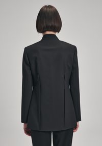 Blazer negro a medida con cuello muesca, corte estructurado y una discreta abertura en la parte trasera. Tejido texturizado, diseño minimalista y mangas largas.