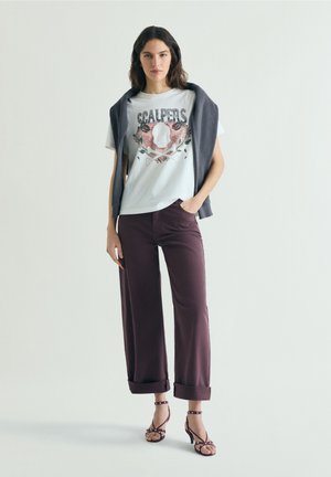 Vrouw staat met hand in zak, draagt een wit T-shirt met schedelprint, donker opgerolde broek, sandalen met bandjes en een trui over de schouders gedrapeerd.