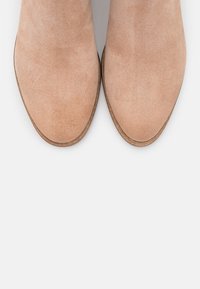 Bottes beiges en daim jusqu'à la cheville, avec un bout arrondi et un talon légèrement surélevé, présentant une texture lisse et des détails de design minimalistes.