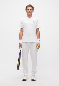 Νέος άνδρας με ολοκληρωμένη λευκή εμφάνιση τένις, κρατώντας ρακέτα στο ένα χέρι και μια μπάλα του τένις στο άλλο, στέκεται μπροστά σε λευκό φόντο.