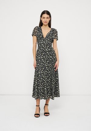 Woman with long dark hair wearing a black midi dress with white daisy print and short puff sleeves, standing against a plain white background.