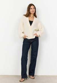 Chunky cream cardigan with a V-neck, ribbed texture, and front buttons. Paired with dark denim jeans and black sandals.