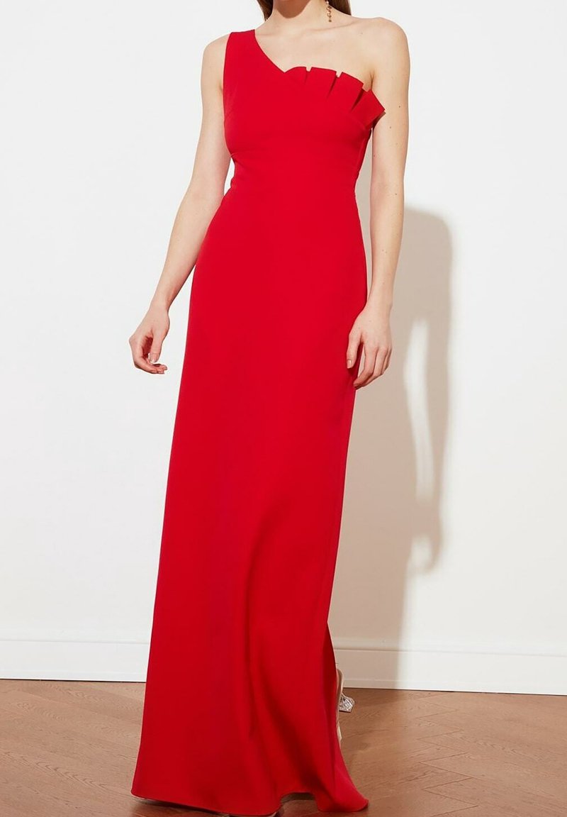 Femme portant une robe rouge longue jusqu'au sol, à une épaule, avec des détails plissés au niveau du décolleté, debout sur un sol en bois contre un mur blanc.