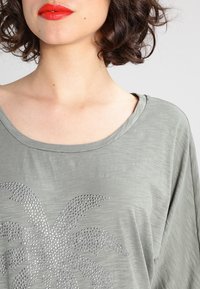 Hellgraues, übergroßes T-Shirt mit weitem Halsausschnitt und kurzen Ärmeln, das auf der Vorderseite ein Design mit einem strassbesetzten Palmenbaum zeigt.