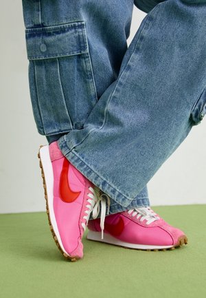 Osoba nosząca niebieskie dżinsy i różowe buty sportowe Nike z czerwonym znakiem, stoi na zielonej podłodze.