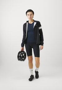 Schwarze Fahrradjacke mit weißen Akzenten, marineblaues Oberteil, schwarze Fahrradshorts und schwarzer Fahrradhelm. Die Schuhe sind schwarz mit Riemen; knielange weiße Socken.