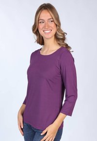 Lila Langarmoberteil mit rundem Ausschnitt und slim Fit; hat eine kleine Brusttasche und einen weichen, glatten Stoff. Getragen mit blauen Jeans.