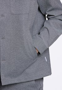 Graues Langarmshirt aus strukturiertem Stoff, mit einer Taschenfront und sichtbaren Nähten. Enthält ein kleines Logoschild am Saum.