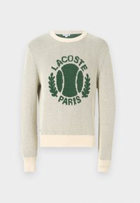 Pulover tricotat de culoare albă cu logo verde și motiv de tenis. Gât rotund, mâneci lungi, manșete și tiv ribbate. Material texturat, ușor.