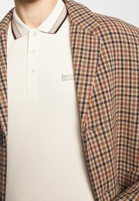 Rutig blazer i brunt, beige och svart med en texturerad yta, kombinerad med en krämfärgad pikétröja med tvåknappsknäppning och präglad logotyp.