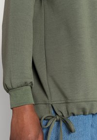 Zbliżenie osoby w oliwkowozielonej bluzie z długim rękawem, z mankietami i ściągaczem na dole, noszącej niebieskie dżinsy.