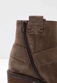 Botín de ante marrón hasta el tobillo con cremallera lateral, detalles de costura cruzada y un tacón apilado texturizado.