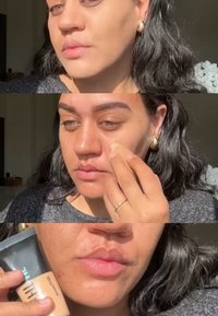 Un tube de fond de teint tenu d'une main, texture crémeuse appliquée sur le visage avec les doigts. Teint clair, cheveux foncés, éclairage doux et pull visible.