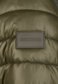 Oliwkowa kurtka puchowa o gładkiej fakturze, z prostokątną, wytłoczoną naszkicowaną metką "HECHTER" przyszytą na kontrastowej łatce.