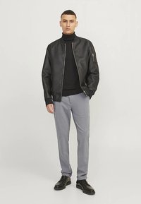 Veste aviateur en cuir noir portée sur un col roulé foncé, associée à un pantalon gris clair et des chaussures noires. Coupe ajustée, texture lisse.