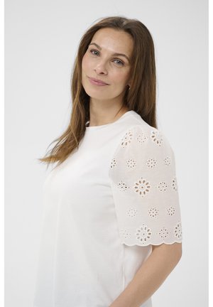 Frau mit glatten braunen Haaren trägt eine weiße Bluse mit kurzen, gemusterten Lochstickärmel und blickt sanft zur Kamera vor einfachem Hintergrund.