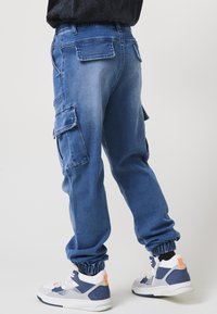 Közepes mosású farmer cargo nadrág rugalmas bokarésszel, több zsebbel és laza szabással. Fehér és kék sportcipővel párosítva.