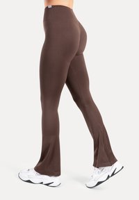 Braune Leggings mit hohem Bund, glatter Textur, figurbetontem Schnitt und ausgestellt am Bein; kombiniert mit weißen Sportschuhen.