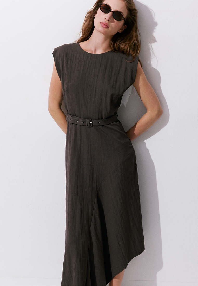 Femme portant une robe midi noire sans manches ceinturée et des lunettes de soleil, debout contre un mur blanc avec les mains dans le dos.