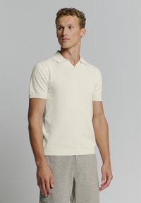Crème korte mouwen polo met een V-hals en geribde manchetten, gemaakt van een zacht materiaal. Draagt bij lichte grijs gemusterde broeken.