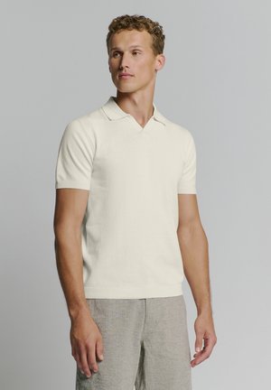 Cream short-sleeve polo shirt with a V-neck and ribbed cuffs, made of a soft material. Worn with light gray patterned trousers.