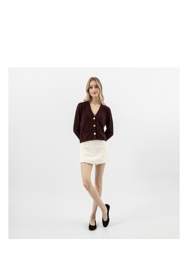 Femme debout, jambes croisées, portant un cardigan bordeaux foncé à gros boutons, une mini-jupe blanche texturée et des ballerines noires sur fond blanc.