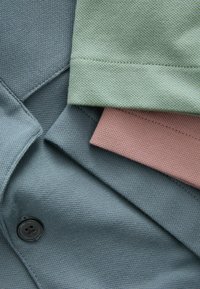 Drei Stoffstücke: eines in dunklem Teal mit Textur und Knopf, eines in hellgrün und eines in gedämpftem Pink, die alle gewebte Muster zeigen.