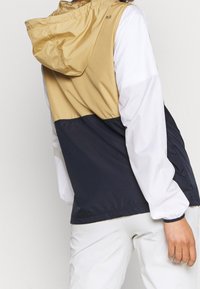 Färgblockerad jacka med en tan övre del, marinblå nedre del och vita ärmar. Tillverkad av lätt material med huva och elastiska manschetter.