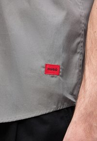 Γκρι ύφασμα με ένα κόκκινο ορθογώνιο επίθεμα με την ένδειξη «HUGO» κοντά στο τελείωμα, δίπλα σε γυμνό χέρι με ορατή τρίχα και ανοιχτόχρωμο δέρμα.