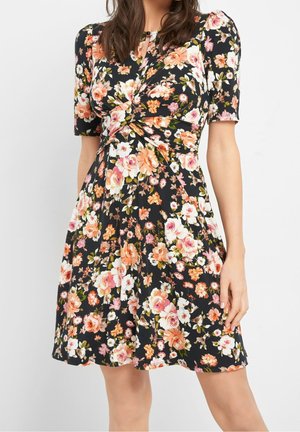 Femme portant une robe noire mi-mollet à imprimé floral rose, blanc et orange, avec des manches courtes bouffantes et un détail de taille plissée.