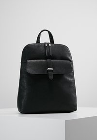 Mochila de cuero negro con asa superior, bolsillo frontal con solapa asegurado por una correa y hebilla, y cierre de doble cremallera sobre una superficie blanca.