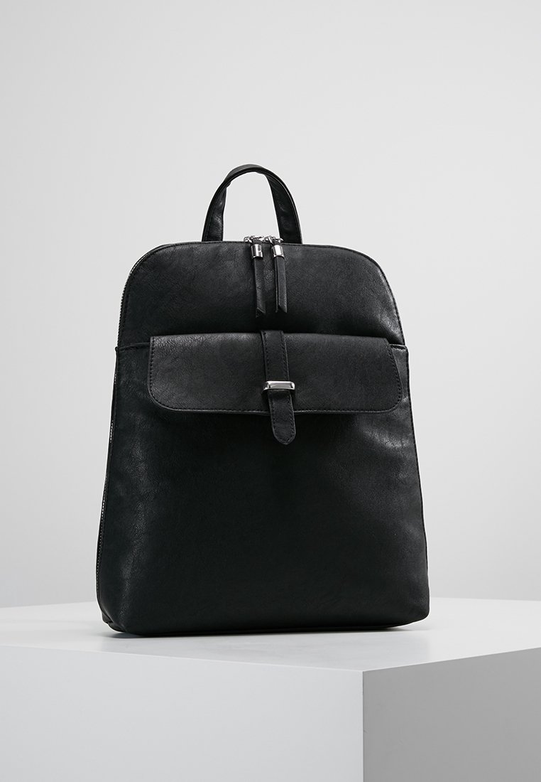 Mochila de cuero negro con asa superior, bolsillo frontal con solapa asegurado por una correa y hebilla, y cierre de doble cremallera sobre una superficie blanca.