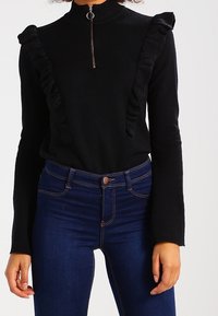 Czarny sweter z podwyższonym kołnierzem i zamkiem z przodu, z falbaniastymi akcentami po obu stronach. Noszony z ciemnoniebieskimi dżinsami o wysokim stanie.