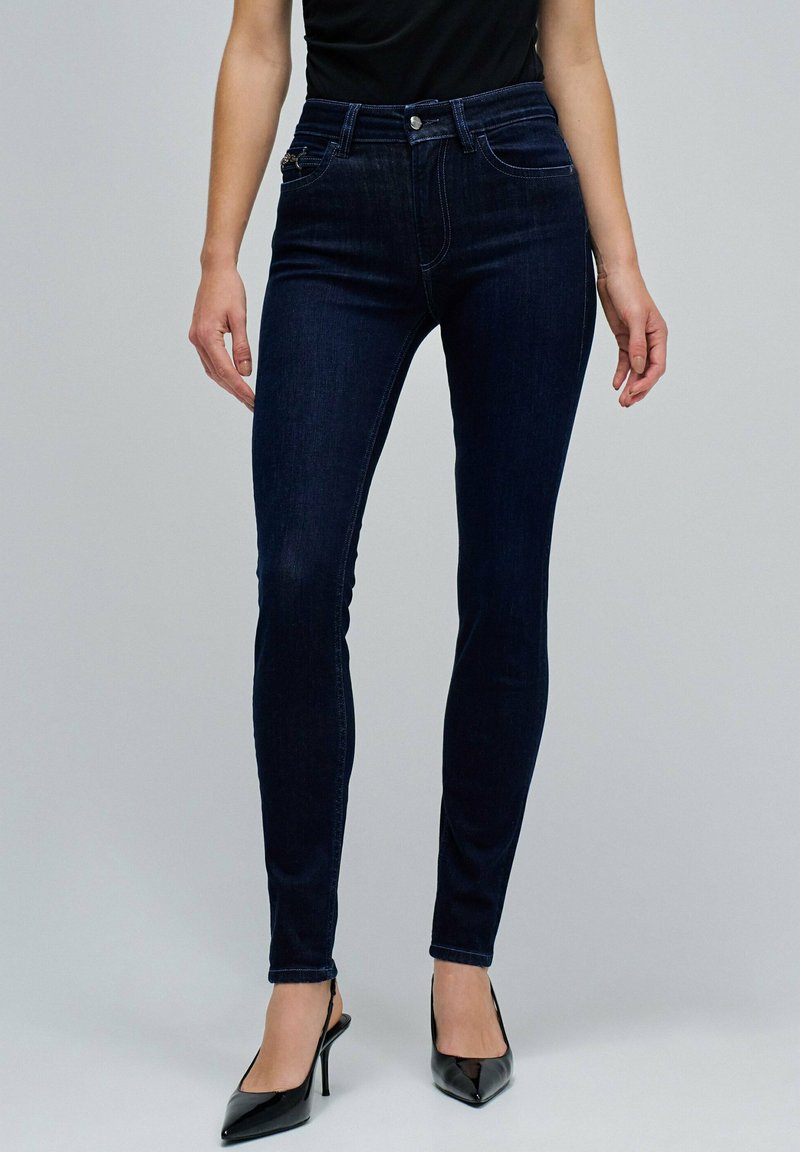 Jeans de mezclilla azul oscuro ajustados con una cintura alta, que presentan bolsillos frontales y una textura suave; combinados con tacones negros de punta.