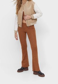 Chaqueta bomber de cuero sintético beige con mangas crema; pantalones acampanados marrones con un patrón ondulado; botas plataforma negras y gruesas.