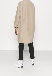 Manteau long beige à coupe droite, poignets boutonnés et fente dans le dos. Associé à un pantalon foncé et des chaussures de sport blanches.